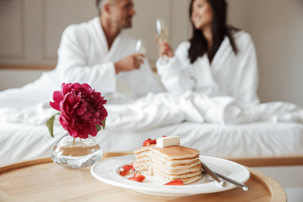 Cropped blurry photo of romantic couple caucasian man and asian woman wearing white housecoat smiling to each other and drinking sparkling wine during breakfast in bed at hotel room - Zdjęcie, obraz