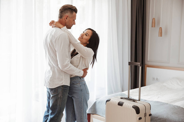 Image of multiethnic couple man and woman wearing casual clothing hugging together in hotel room with big luggage. Vacation concept - Foto, afbeelding