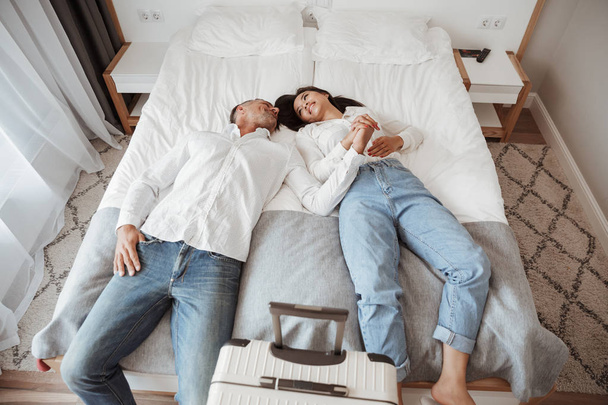 Photo from above of joyful couple man and woman in casual jeans clothing resting together and lying in bed at hotel room with big luggage. Holiday or vacation concept - Фото, зображення