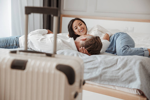 Photo of happy couple man and woman in casual clothing resting together and lying in bed at hotel room with luggage near bed. Holiday or vacation concept - Foto, Imagen