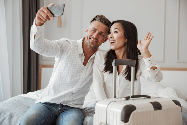 Happy young couple taking selfie with mobile phone while sitting together on bed at a hotel room with a suitcase - Photo, Image