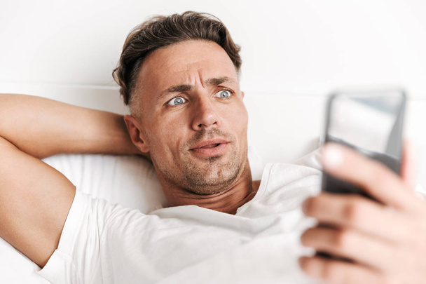 Close up of shocked man using mobile phone while laying in bed - Φωτογραφία, εικόνα