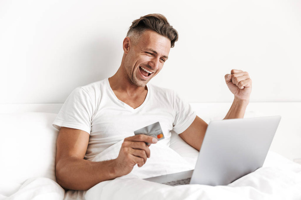 Satisfied man using laptop computer and celebrating success while sitting in bed and holding credit card - Фото, зображення