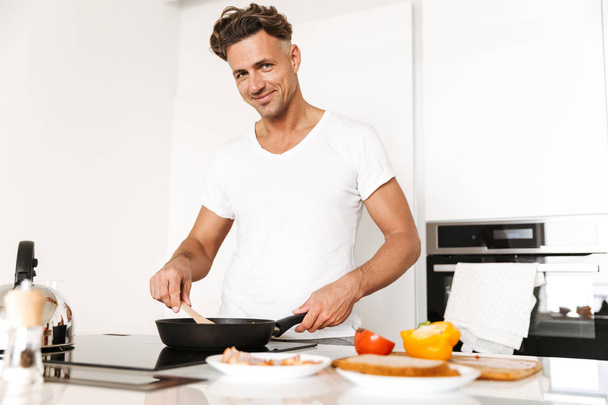 Attractive man cooking eggs for breakfast while standing at a kitchen - Фото, изображение