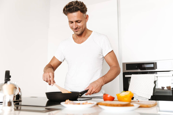 Smiling man cooking eggs for breakfast while standing at a kitchen - Foto, afbeelding