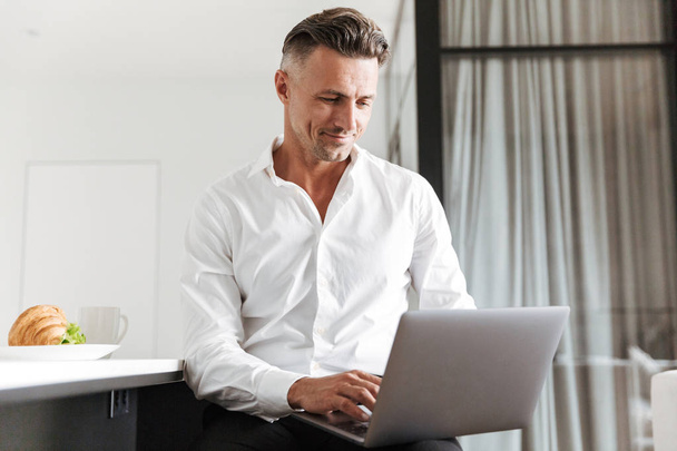 Smiling man dressed in formal clothes using laptop while sitting at home and having breakfast - Foto, Bild