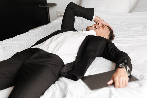 Desperate man dressed in suit lying on bed with arms at his face holding laptop - Фото, изображение
