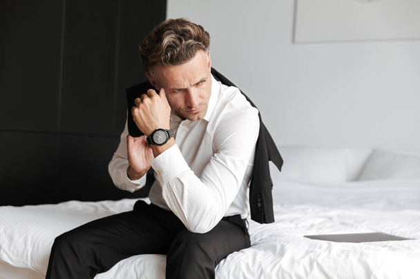 Depressed man dressed in suit sitting on bed and looking away - Foto, afbeelding