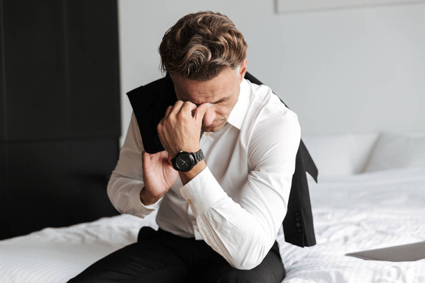 Stressed man dressed in suit sitting on bed and looking down - Fotó, kép