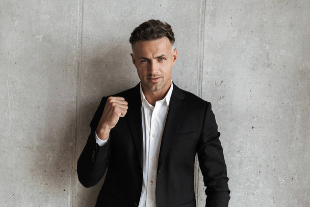 Handsome man dressed in suit showing his fist and looking at camera over gray wall background - Foto, imagen
