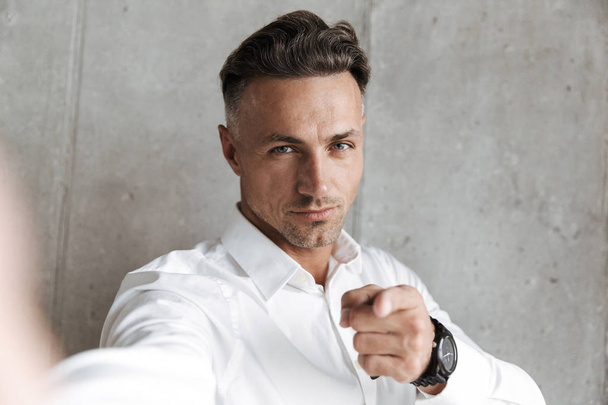 Portrait of a confident man in white shirt taking a selfie with outsretched hand and pointing at camera over gray wall background - Фото, зображення