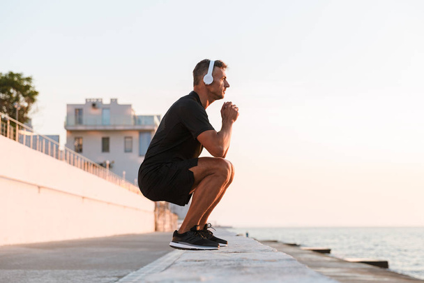 Photo of healthy caucasian man 30s in tracksuit warming up and squatting off coast while listening to music via wireless headphones during sunrise - Foto, imagen
