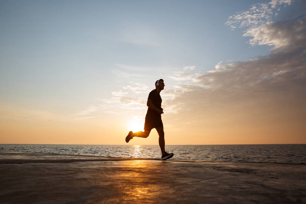 Silhouette of young man in sportive shorts and t-shirt running along pier offshore and listening to music via wireless earphones during sunrise - Photo, image