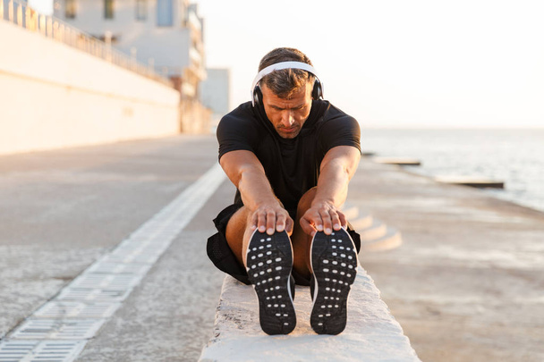 Photo of healthy caucasian man 30s in tracksuit warming up and stretching legs off coast while listening to music via wireless headphones during sunrise - 写真・画像