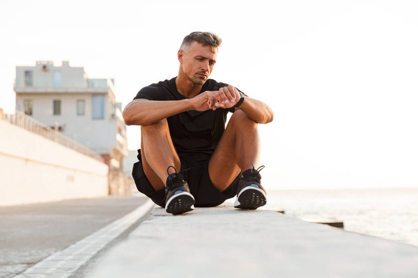 Photo of handsome caucasian man 30s in tracksuit sitting on parapet of boardwalk near seaside and looking at wrist smart watch - Фото, изображение