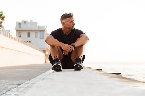 Photo of concentrated strong man 30s in tracksuit sitting on parapet of boardwalk near seaside during sunrise - Фото, изображение