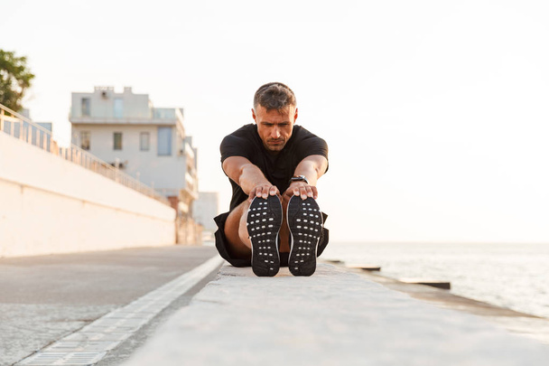 Image of caucasian sportsman 30s in tracksuit sitting on parapet and stretching legs near seaside during summer morning - Photo, image