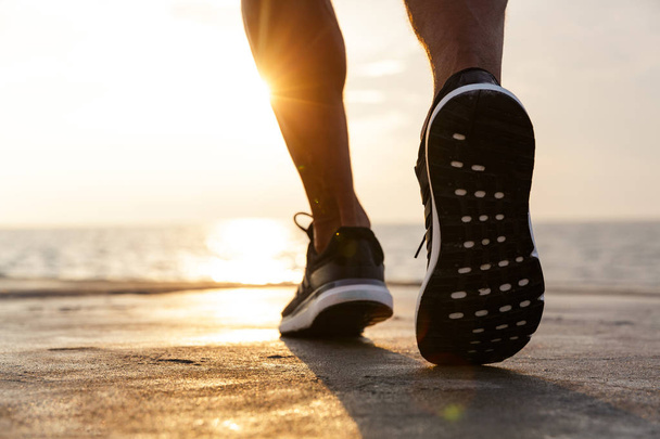 Image of sunlit caucasian athlete guy wearing sneakers running along pier at seaside during beautiful sunrise - Φωτογραφία, εικόνα