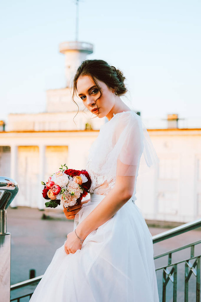 Adorable caucasian bride in white flowing wedding dress made of lace and tulle standing on the staircase in sunset evening - Fotografie, Obrázek