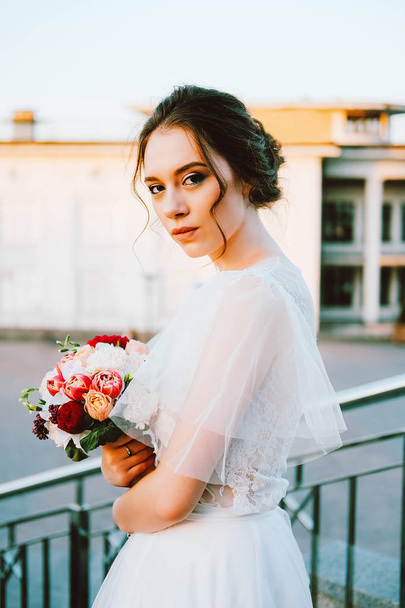Adorable caucasian bride in white flowing wedding dress made of lace and tulle standing on the staircase in sunset evening - Zdjęcie, obraz