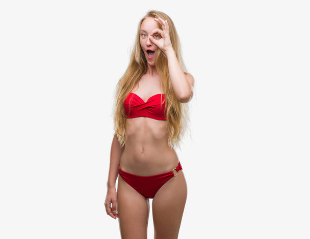Adolescente blonde portant un bikini rouge faisant un geste correct choquée par le visage surpris, les yeux regardant à travers les doigts. Expression incrédule
. - Photo, image