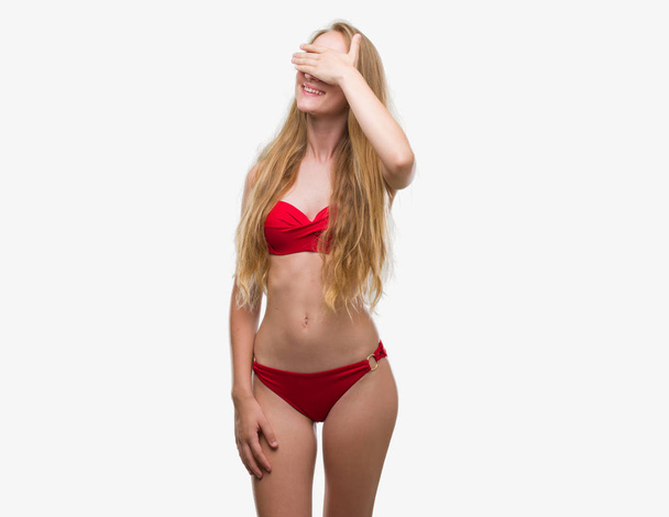 Szőke tizenéves nő visel piros bikini mosolyogva és nevetve a kezét, arcát, szemét, meglepetés, amely. Vak koncepció. - Fotó, kép