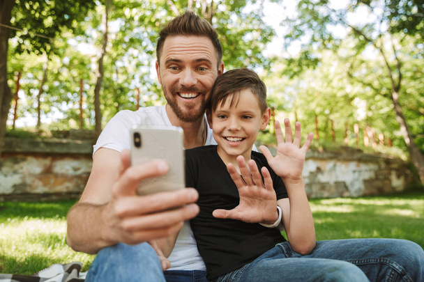 Image of happy young father sitting with his little son outdoors in park nature using mobile phone waving to family or friends. - Photo, Image