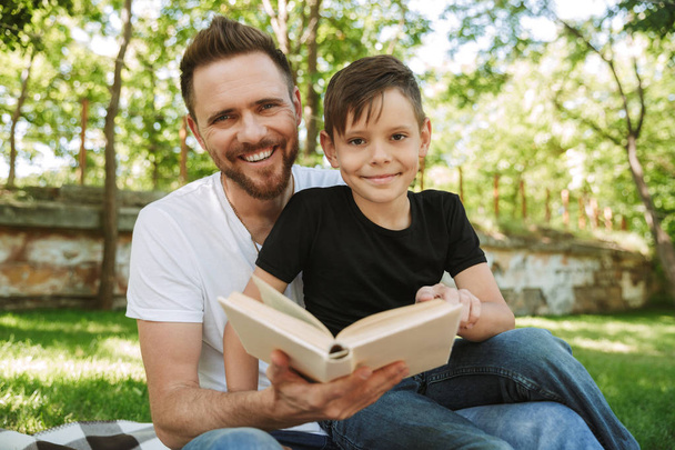 Image of happy young father sitting with his little son outdoors in park nature reading book. - Fotografie, Obrázek