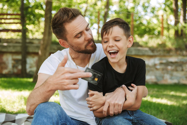 Photo of happy young father sitting with his little son outdoors in park nature holding credit card. - Photo, image