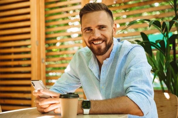 Photo of happy young bearded man in cafe outdoors using chatting by mobile phone drinking coffee looking camera. - Foto, Bild