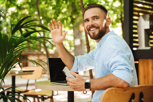 Photo of smiling emotional young bearded man outdoors using laptop computer and mobile phone waving to friends. - Foto, afbeelding