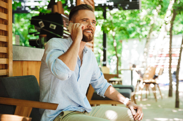 Photo of handsome happy young bearded man outdoors looking aside talking by mobile phone. - Fotoğraf, Görsel