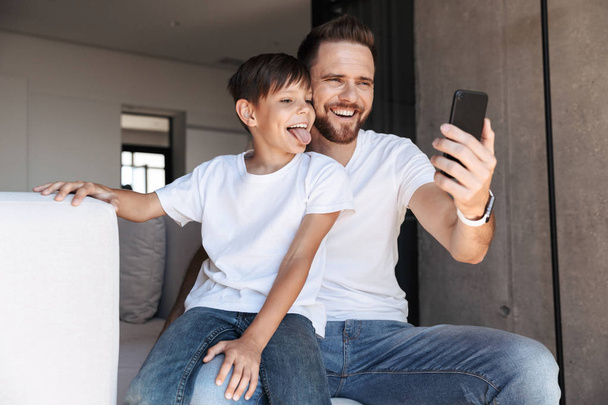 Image of cheerful young man father dad using mobile phone with his son indoors at home. - Foto, Imagen