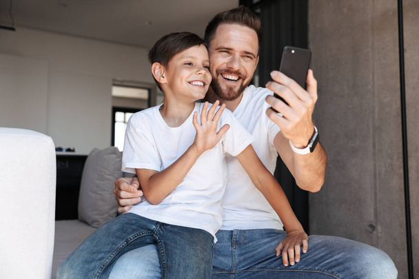 Image of handsome bearded man indoors at home with his son child using mobile phone waving. - Fotografie, Obrázek