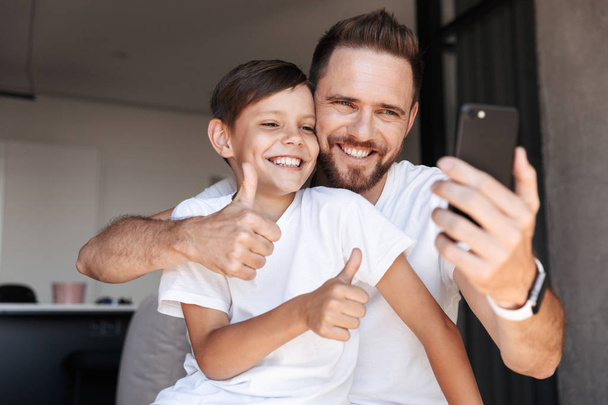 Image of handsome bearded man indoors at home with his son child using mobile phone make thumbs up gesture take a selfie. - Fotoğraf, Görsel