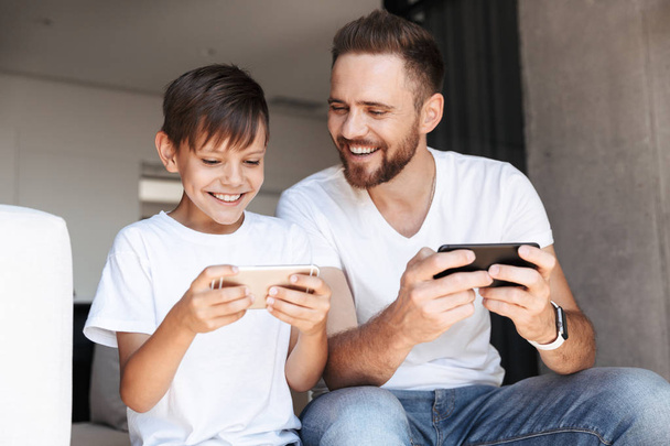 Image of handsome bearded man indoors at home with his son child play games by mobile phones. - Foto, Imagem