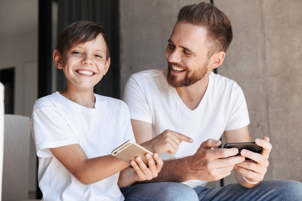 Image of handsome bearded man indoors at home with his son child play games by mobile phone. Boy pointing to father's phone looking camera. - Photo, image