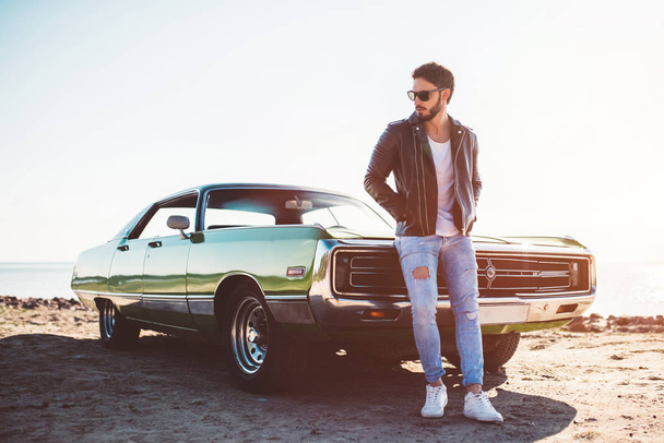 Handsome bearded man is standing near his green retro car on the beach. Vintage classic car. - Fotografie, Obrázek