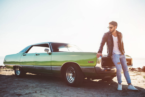 Handsome bearded man is standing near his green retro car on the beach. Vintage classic car. - Foto, Bild