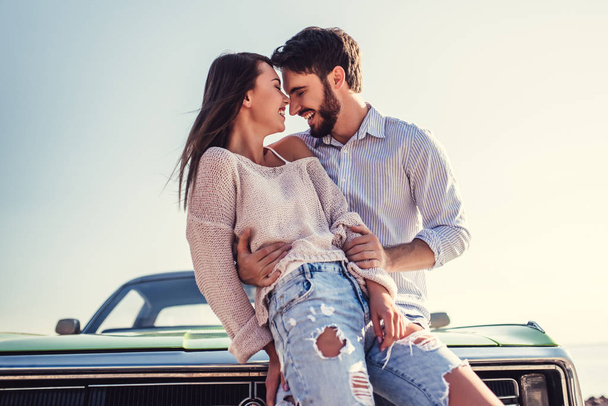 Romantic couple is standing near green retro car on the beach. Handsome bearded man and attractive young woman with vintage classic car. Love story. - Zdjęcie, obraz