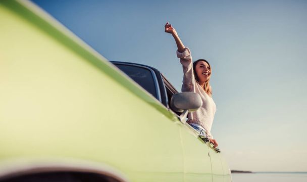 Feel the spirit of adventure! Attractive young woman is leaning out of the window of green retro car. Vintage classic car. - Photo, Image