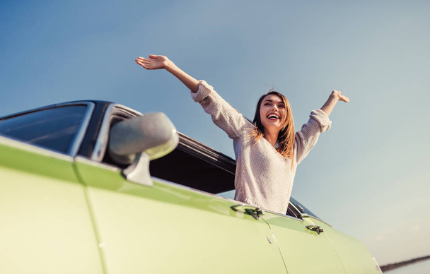 Feel the spirit of adventure! Attractive young woman is leaning out of the window of green retro car. Vintage classic car. - Photo, image