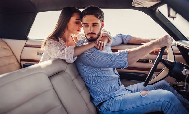 Romantic couple is sitting in green retro car on the beach. Handsome bearded man and attractive young woman in vintage classic car. Stylish love story. Hugging and kissing while being in car. - Foto, Bild