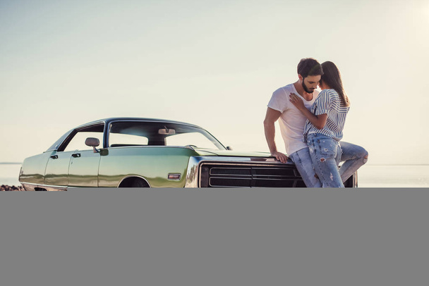 Romantic couple is standing near green retro car on the beach. Handsome bearded man and attractive young woman with vintage classic car. Love story. - 写真・画像