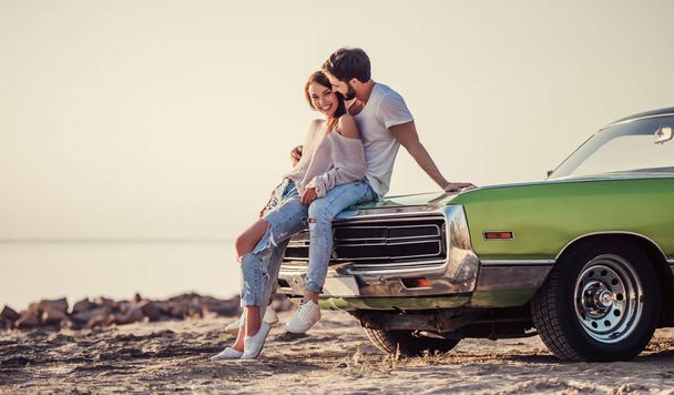 Romantic couple is standing near green retro car on the beach. Handsome bearded man and attractive young woman with vintage classic car. Love story. - Фото, изображение