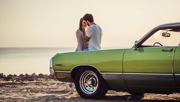Romantic couple is standing near green retro car on the beach. Handsome bearded man and attractive young woman with vintage classic car. Love story. - Foto, Imagen