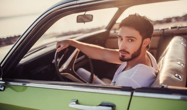 Handsome bearded man is sitting in his green retro car standing on the beach. Vintage classic car. - Foto, immagini
