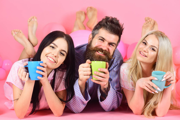 Man and women in domestic clothes, pajamas. Lovers drinking coffee in bed. Lovers in bed concept. Man and women, friends on happy faces lay, pink background. Threesome relax in morning with coffee - Photo, image