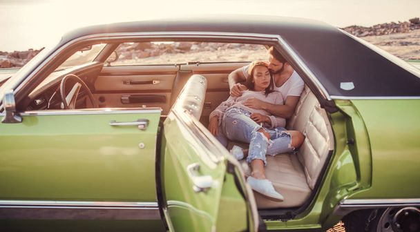 Romantic couple is sitting in green retro car on the beach. Handsome bearded man and attractive young woman in vintage classic car. Stylish love story. Hugging and kissing while being in car. - Photo, Image