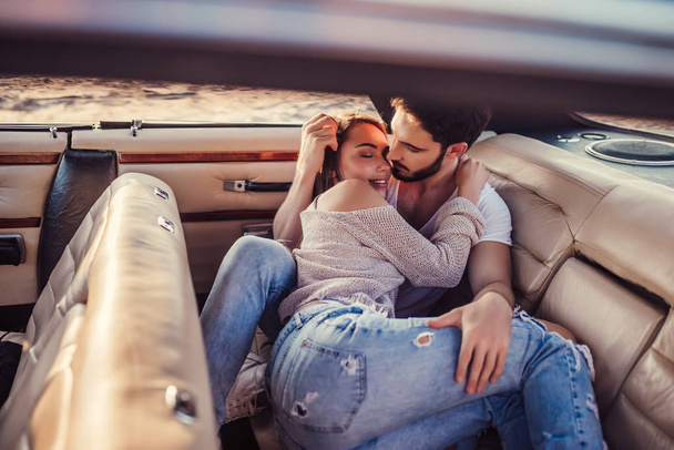 Romantic couple is sitting in green retro car on the beach. Handsome bearded man and attractive young woman in vintage classic car. Stylish love story. Hugging and kissing while being in car. - Zdjęcie, obraz